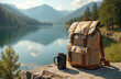 Lightweight brown backpack with black cup rests on rock near tranquil lake. Natural background mountains hills forest. Summer hiking camping trip adventure. Concept travel tourism expedition journey