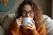 © Viktor - Smiling woman enjoys hot tea in cozy home environment. Girl wears glasses, comfy sweater. Relaxing at home on sofa. Perfect for winter vibes, autumn season, leisure activity.