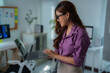 © MINAE - Young, beautiful Asian businesswoman wearing glasses and a stylish purple shirt, standing confidently while using a laptop in a modern office adorned with informative charts on the wall
