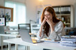© Wasana - A woman is sitting at a desk with a laptop and a stack of papers