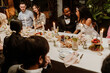 © Drobot Dean - A Black bride in her 20s in a white gown talking with a Black groom in his 20s in a black suit sit with multiethnic guests at a long dining table during the wedding, in a decorated plant-filled venue.