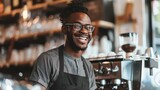 Cheerful young African American barista wearing apron and eyeglasses standing in coffee shop, leaning on counter next to coffee machine, looking away