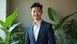 © miss irine - Asian man in suit, smiling, office, indoor plants background. Young businessman in formal attire. Pro corporate employee looks at camera. Confident man portrait.