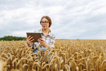 Fototapete - Close-up of a young female farmer with a modern digital tablet checking the quality of the wheat crop outdoors. Handsome agronomist uses a tablet in an agricultural field. Rich harvest concept.