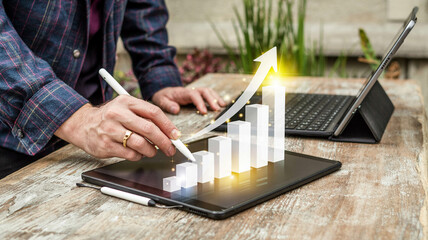 Poster - Person using stylus on tablet with growth chart and laptop on wooden table in outdoor setting view