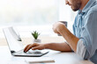 © Prostock-studio - Handsome male freelancer browsing social media on laptop and drinking morning coffee, sitting at desk by the window in home office, enjoying remote work and online distance job, side view, copy space