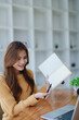 © Jirapong - Smiling young woman engaged in a joyful online e-learning video call, gestu while holding a book with headphones on a wooden table nearby