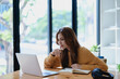 © Jirapong - Smiling young woman engaged in a joyful online e-learning video call, gestu while holding a book with headphones on a wooden table nearby