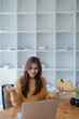 © Jirapong - Smiling young woman engaged in a joyful online e-learning video call, gestu while holding a book with headphones on a wooden table nearby
