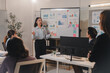 © Phimwilai - Young businesswoman presenting marketing data on whiteboard to team of colleagues listening attentively during a business meeting in modern office