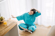 © irissca - Young white woman in a turquoise robe does a neck stretch while sitting cross-legged on a mat in a bright room. Soft wellness, mindful morning routine, home self-care, body awareness