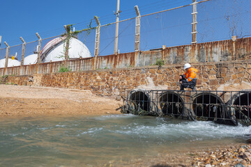 an environmental engineer in safety gear sits on a concrete drainage outlet near a factory, observin