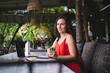 © BullRun - Woman in red dress holding a coffee cup, looking thoughtful while seated at a laptop in an outdoor workspace with lush greenery. Relaxed, creative setting with natural decor and bohemian vibe