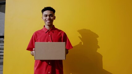 Aufkleber - Young smiling delivery man in red shirt is holding a cardboard box against yellow background, representing successful and fast shipping service