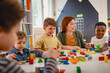 © lordn - Preschool teacher interacts with a diverse group of children building with colorful blocks in a bright classroom, promoting creativity, teamwork, and multicultural early education.