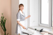 © Pixel-Shot - Smiling maid with hand duster cleaning table near window in living room