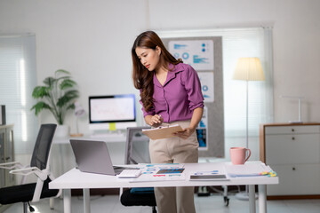Poster - Asian businesswoman standing near desk reviewing documents and working with laptop in office