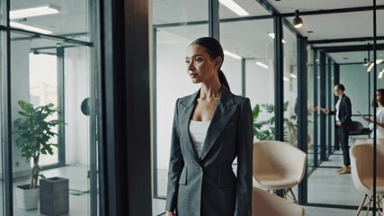 Aufkleber - Confident businesswoman stands near window, contemplating as colleagues engage in discussion in background, showcasing focus and leadership in modern office environment