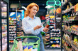 © Home-stock - Positive woman scanning qr code of product, holding glass jar and using smartphone, standing near shelves. Concept of shopping and consumerism.