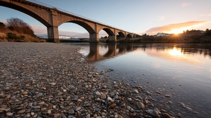 Aufkleber - A tranquil evening unfolds as the sun sets, casting warm hues over a serene river. Soft reflections ripple beneath an ancient stone bridge, inviting calm thoughts