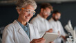 © Sajib - Woman in lab coat looking at tablet with colleagues in background performing research work in lab