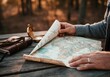 © SERHII - Person unfolding vintage world map on wooden table in forest setting at sunset. Analog Travel Concept