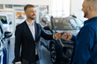 © Anastasiya - Smiling salesman giving car keys to male customer in bright modern car dealership showroom, auto salon manager in black suit greeting man with purchase, happy man buying vehicle