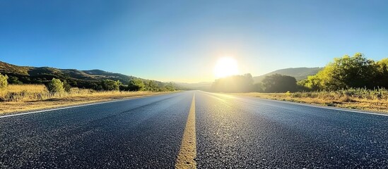  Asphalt road under bright sunlight stretching toward distant hills, green fields on both sides with scattered trees, wide-angle composition with gradient blue sky, serene rural landscape.