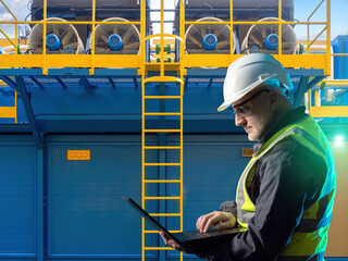 Poster - Engineer in safety gear uses a laptop to monitor or configure industrial ventilation or cooling systems, working in the field of industrial maintenance and engineering.