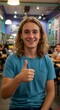 © Gabriel - Smiling young man with long blond hair gives a thumbs up in a brightly lit cafe setting, positive expression.