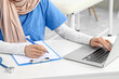© Pixel-Shot - Female Muslim doctor working with laptop and clipboard at table in clinic, closeup