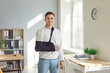 © Studio Romantic - Portrait of a young smiling man standing at home or at office on rehabilitation with his arm in a plaster cast after injury. Patient wearing sling or bandage. Recovery and rehab concept.