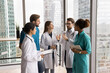 © fizkes - Informal brainstorming. Focused multiethnic doctors interns team stand in clinic office with glass walls engaged in open communication listen to young physician teammate expressing idea for discussion