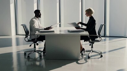 Poster - Professional business colleagues discussing project details while sitting at white table in bright, modern office environment, exchanging documents and collaborating with focused interaction