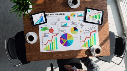 Poster - Businesswoman sitting at desk analyzing business strategy on a large sheet of paper with colorful charts, graphs, and data, using digital tablets for additional information, overhead view