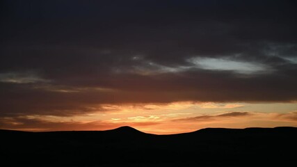 Wall Mural - time lapse of evening orange colored clouds after sunset above rolling hills silhouetted