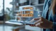 © Usama - Person using a tablet to visualize a holographic model of a modern building, representing digital real estate business.