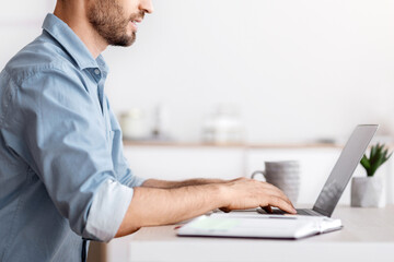 Poster - Handsome millennial businessman sitting at workplace in white office, working on laptop, typing on computer keyboard, answering emails, corresponding with clients, side view with copy space