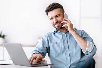 Poster - Cheerful Young Man Working Remotely From Home Office With Cellphone And Laptop, Sitting At Desk In Room, Copy Space