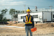 © ultramansk - Male worker looks at construction plan with construction work behind him at roof truss construction site under construction. Workers in safety gear carry out inspection and repair work.