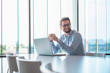 Smiling businessman with laptop and coffee in modern office