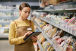 © Seventyfour - Employee in grocery store using digital tablet while checking inventory on shelves, wearing an apron with name badge. Shelves stocked with various products in the background