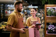© Seventyfour - Two grocery store employees smiling and conversing while handling store tasks. Male and female staff members holding a cardboard box and tablet in the bustling supermarket environment