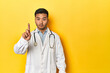 © Asier - Asian doctor in white coat with stethoscope, yellow studio showing number one with finger.
