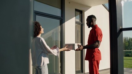 Aufkleber - Delivery man in bright red uniform handing package to smiling woman standing at contemporary home's front entrance during daytime sunlight