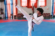 © Studio Marmellata - A male martial artist in a white uniform and black belt performs a high side kick on a blue mat inside a taekwondo gym with red curtains and chairs in the background