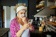 © Marko Geber - Worried senior woman with face mask looking at smartphone in kitchen