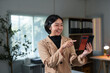 © amnaj - Young Asian businesswoman in smart casual attire having a video call on smartphone in office, pointing at screen and smiling