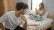 © Krakenimages.com - Man sitting on bed looking at woman holding phone in cozy bedroom setting indoors, suggesting a couple scenario in a modern home environment.