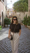 © Krakenimages.com - Woman smiling on a cobblestone street in rome, italy, wearing sunglasses and beige pants, with surrounding old town architecture and greenery under sunny skies.